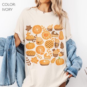 May include: Ivory t-shirt with a fall-themed design. The graphic features pumpkins, pies, cupcakes, and autumn leaves in orange, brown, and yellow. The text "COLOR: IVORY" is at the top left of the image.