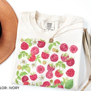 Op de afbeelding: Ivoor kleurig T-shirt met een frambozen ontwerp. Het shirt heeft een ronde hals en korte mouwen. Het frambozenpatroon bevat rode bessen, groene bladeren en witte bloemen. Het shirt is gelabeld "Comfort Colors" en is maat S.