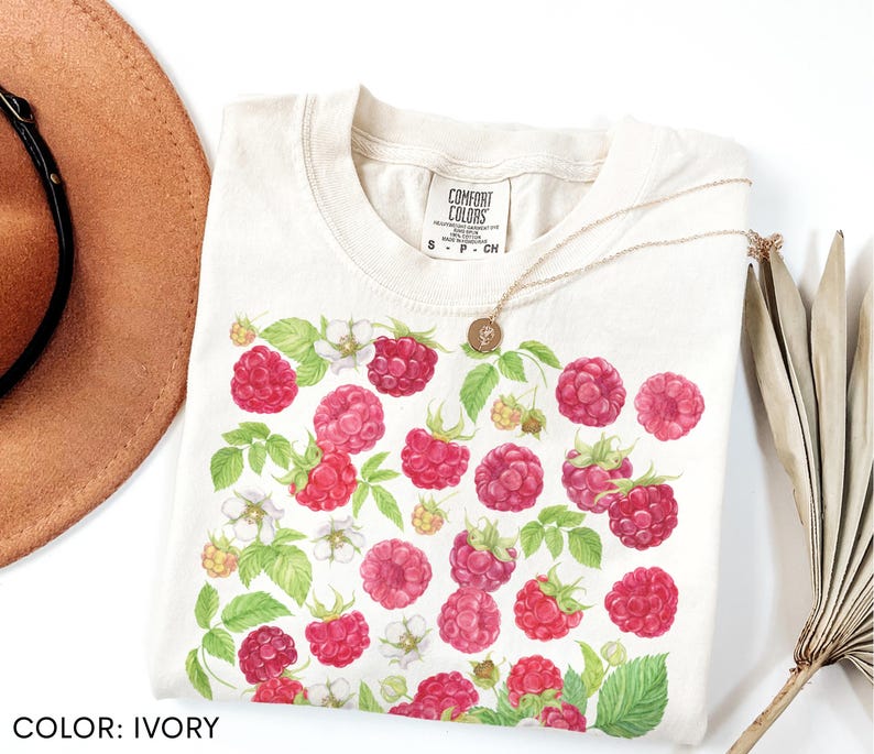 Op de afbeelding: Ivoor kleurig T-shirt met een frambozen ontwerp. Het shirt heeft een ronde hals en korte mouwen. Het frambozenpatroon bevat rode bessen, groene bladeren en witte bloemen. Het shirt is gelabeld "Comfort Colors" en is maat S.