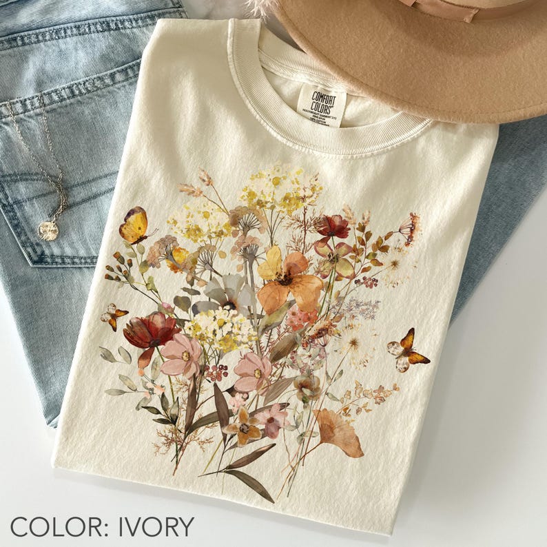 May include: Ivory-colored t-shirt featuring a watercolor floral design with butterflies. The design includes flowers in shades of orange, red, yellow, and pink. The shirt is folded and placed on top of blue jeans and a tan hat.