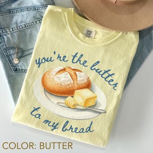 May include: A butter-colored t-shirt with the phrase "you're the butter to my bread" in blue script. The graphic features a watercolor illustration of a bread roll, butter, and a knife on a plate. The shirt is folded on top of blue jeans.