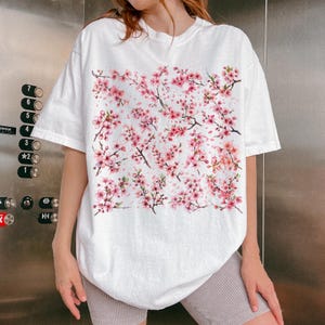 May include: White t-shirt featuring a watercolor design of pink cherry blossom branches and flowers. The short-sleeved shirt has a relaxed fit. The floral pattern covers the front of the shirt, creating a spring-themed aesthetic.