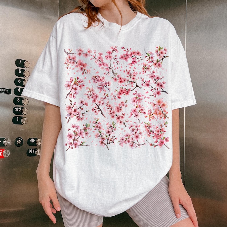 May include: White t-shirt featuring a watercolor design of pink cherry blossom branches and flowers. The short-sleeved shirt has a relaxed fit. The floral pattern covers the front of the shirt, creating a spring-themed aesthetic.