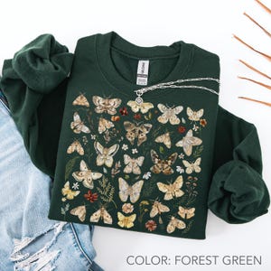 May include: A forest green crewneck sweatshirt featuring a detailed graphic print of various moths and small wildflowers in shades of brown, beige, and cream. The garment has long sleeves and a ribbed collar with a visible brand tag at the neckline.