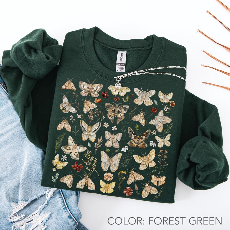 May include: A forest green crewneck sweatshirt featuring a detailed graphic print of various moths and small wildflowers in shades of brown, beige, and cream. The garment has long sleeves and a ribbed collar with a visible brand tag at the neckline.
