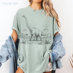 Pu&ograve; includere: T-shirt verde chiaro con un'illustrazione in bianco e nero di un dinosauro Brachiosaurus in un paesaggio preistorico con montagne e palme. Il testo "COLOR: BAY" &egrave; nell'angolo in alto a sinistra.