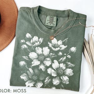 以下が含まれることがあります： モスグリーンのTシャツに白い花の模様。Tシャツはクルーネックで半袖です。ペンダント付きのゴールドネックレスがTシャツの上にあります。「COLOR: MOSS」の文字が画像の下部にあります。