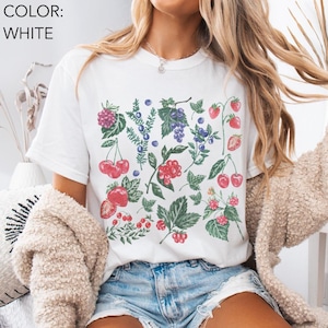 May include: White t-shirt with a vibrant botanical print of cherries, strawberries, raspberries, and blueberries. The shirt has a crew neck and short sleeves. The text "COLOR: WHITE" is in the upper left corner.