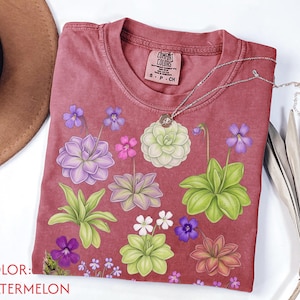 May include: A watermelon-coloured t-shirt featuring a graphic print of various succulent and flowering plants in shades of purple, green, and pink. The shirt has a crew neckline and a Comfort Colors brand label on the inside collar, size S-CH.