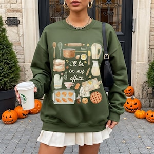 Peut inclure: Sweat-shirt vert olive avec un motif sur le thème de la pâtisserie. Le motif comprend des ustensiles de cuisine, des pâtisseries et le texte "I'll be in my office". La tenue est complétée par une jupe plissée blanche.