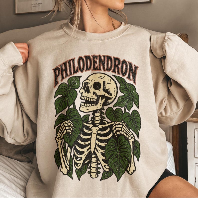 May include: Beige sweatshirt featuring a graphic of a skeleton surrounded by green leaves. The word "PHILODENDRON" is arched across the top in a brown and green font. The design is printed on a neutral-colored, long-sleeved sweatshirt.
