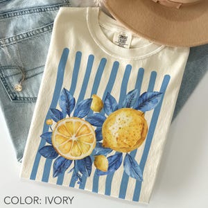 Ivory Comfort Colors t-shirt with blue toile-inspired stripes and watercolor lemons, featuring soft blue leaves in a coquette cottagecore aesthetic, styled in a flat lay with denim and accessories