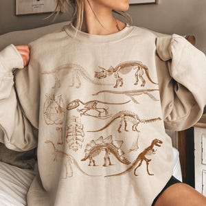 Op de afbeelding: Een beige sweater met ronde hals en een bruine grafische print van diverse dinosaurusskeletten, waaronder een triceratops, een stegosaurus en een tyrannosaurus rex, gerangschikt in een wetenschappelijke illustratiestijl op de voorzijde.
