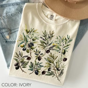 May include: Ivory-colored t-shirt featuring a watercolor design of olive branches with green leaves and black olives. The shirt has a crew neck and the brand "Comfort Colors" is visible. The text "COLOR: IVORY" is at the bottom.