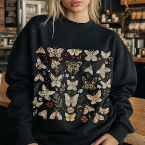 May include: A black crewneck sweatshirt featuring a detailed graphic print of various moths and butterflies in shades of beige, brown, and cream, interspersed with small botanical elements like leaves and tiny flowers across the front chest area.