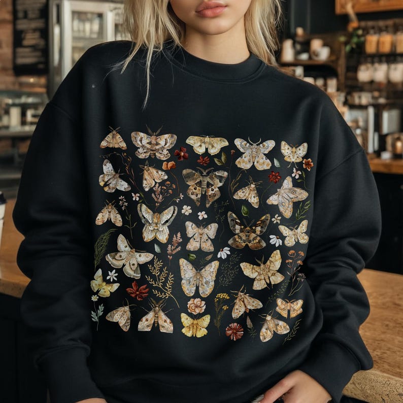 May include: A black crewneck sweatshirt featuring a detailed graphic print of various moths and butterflies in shades of beige, brown, and cream, interspersed with small botanical elements like leaves and tiny flowers across the front chest area.
