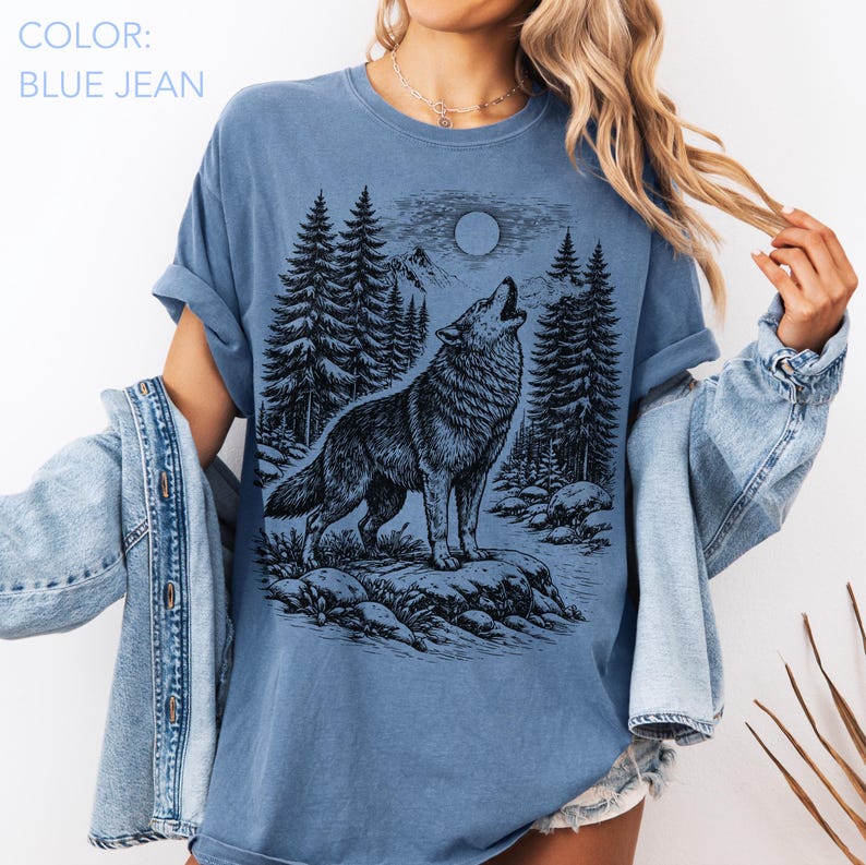 Puede incluir: Una camiseta de color azul vaquero con un estampado gr&aacute;fico negro de un lobo aullando a la luna en un entorno forestal con pinos y terreno rocoso. La camiseta tiene cuello redondo y mangas cortas, combinada con una chaqueta de mezclilla.