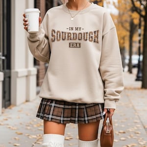 May include: Beige sweatshirt with the text "-IN MY- SOURDOUGH ERA" in brown and tan lettering. The person is wearing a brown plaid pleated skirt and white knee-high socks. A brown purse and a white coffee cup are also visible.