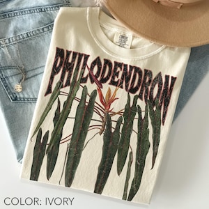 May include: An ivory t-shirt featuring a graphic print of dark green philodendron leaves and a central orange flower. The word PHILODENDRON is printed in large, distressed black capital letters above the botanical illustration. The shirt has a crew neckline.