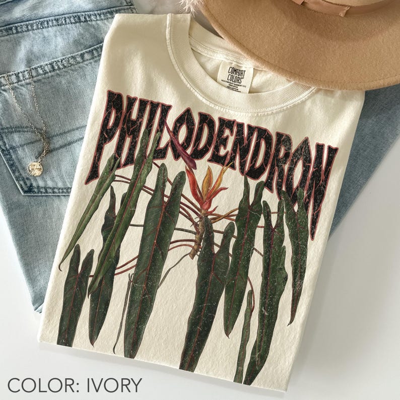 May include: An ivory t-shirt featuring a graphic print of dark green philodendron leaves and a central orange flower. The word PHILODENDRON is printed in large, distressed black capital letters above the botanical illustration. The shirt has a crew neckline.