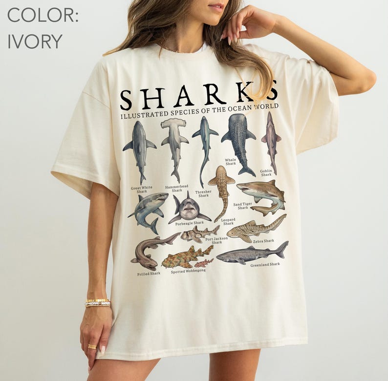 May include: An ivory short-sleeve t-shirt featuring a graphic print titled SHARKS ILLUSTRATED SPECIES OF THE OCEAN WORLD. The design displays various shark species including the Great White, Hammerhead, Thresher, Whale, Goblin, Porbeagle, Leopard, Sand Tiger, Frilled, Port Jackson, Zebra, Spotted Wobbegong, and Greenland shark.