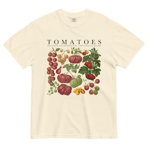 May include: Cream-colored t-shirt featuring a botanical illustration of various tomatoes. The design includes red, yellow, and green tomatoes, with the word "TOMATOES" and "SOLANUM LYCOPERSICUM - BOTANICAL STUDY" printed above the illustration.