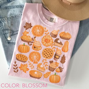May include: A light pink t-shirt with a fall-themed design featuring pumpkins, pies, leaves, and mugs. The shirt has a crew neck and the text "COLOR: BLOSSOM" at the bottom. A tan hat and blue jeans are in the background.