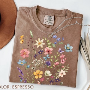 Puede incluir: Camiseta color espresso con un dise&ntilde;o floral de flores rosas, azules, amarillas y moradas. La camiseta tiene cuello redondo y mangas cortas. Un collar plateado con un colgante est&aacute; sobre la camiseta.
