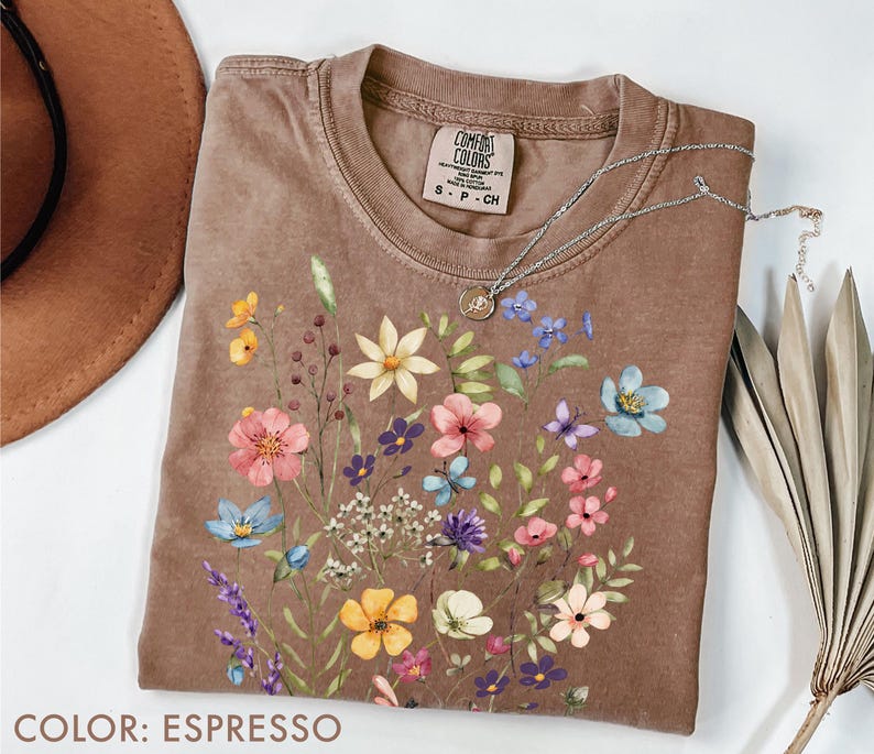 Puede incluir: Camiseta color espresso con un dise&ntilde;o floral de flores rosas, azules, amarillas y moradas. La camiseta tiene cuello redondo y mangas cortas. Un collar plateado con un colgante est&aacute; sobre la camiseta.