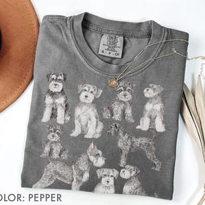 Puede incluir: Camiseta color pimienta con ilustraciones de perros Schnauzer. La camiseta tiene cuello redondo y mangas cortas. Un collar plateado con un colgante está drapeado sobre la camiseta. El texto "COLOR: PEPPER" está en la parte inferior.