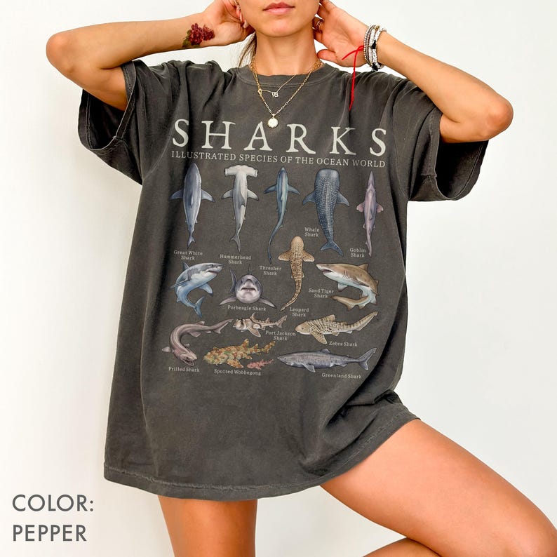 May include: A pepper-colored oversized t-shirt featuring a graphic print titled SHARKS ILLUSTRATED SPECIES OF THE OCEAN WORLD. The design displays various shark species with their names printed underneath, including Great White, Hammerhead, and Whale Shark.