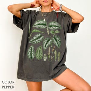 以下が含まれることがあります： ペッパーカラーのTシャツで、緑と銀の斑入りの葉や植物の構造を描いた植物画がプリントされています。グラフィックには、複数のハート型の葉の模様と小さな植物のつぼみが中央に配置されています。