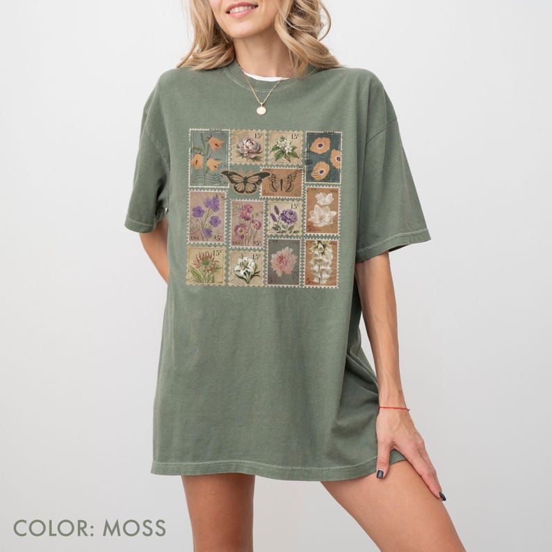 K&ouml;nnte beinhalten: Moosgr&uuml;nes T-Shirt mit einem Aufdruck von Vintage-Briefmarken. Die Briefmarken zeigen Blumen- und Schmetterlingsdesigns in Orange-, Lila- und Rosat&ouml;nen. Das T-Shirt hat kurze &Auml;rmel und eine lockere Passform. Der Text "COLOR: MOSS" steht unten.