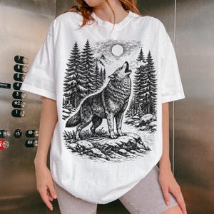 Puede incluir: Una camiseta blanca de manga corta con una ilustraci&oacute;n gr&aacute;fica en blanco y negro de un lobo aullando a la luna llena en un entorno forestal con pinos, terreno rocoso y monta&ntilde;as al fondo, representada en un estilo de boceto detallado.