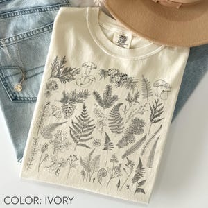 May include: Ivory-colored t-shirt with a crew neck, featuring a detailed black and white botanical illustration of various plants, mushrooms, and ferns. The text "COLOR: IVORY" is at the bottom. The shirt is folded and placed on a pair of blue jeans.