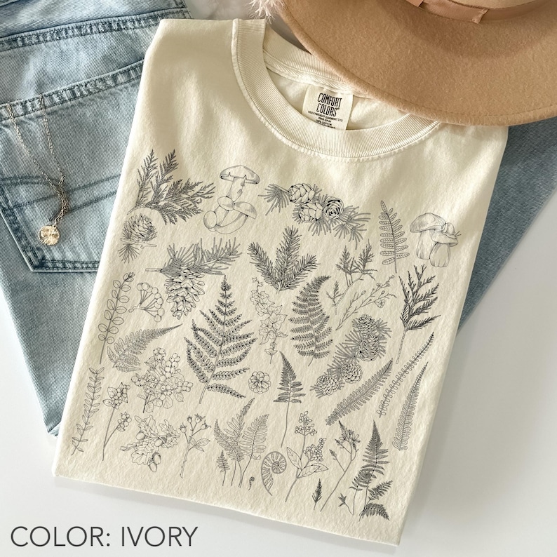 May include: Ivory-colored t-shirt with a crew neck, featuring a detailed black and white botanical illustration of various plants, mushrooms, and ferns. The text "COLOR: IVORY" is at the bottom. The shirt is folded and placed on a pair of blue jeans.