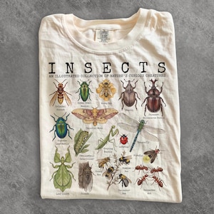 Puede incluir: Una camiseta de color crema con un estampado gráfico titulado INSECTS AN ILLUSTRATED COLLECTION OF NATURE'S CURIOUS CREATURES. El diseño muestra ilustraciones detalladas de varios insectos, incluyendo un avispón, un escarabajo, una polilla y una mantis.