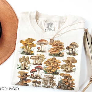 Pu&ograve; includere: T-shirt color avorio con un'illustrazione dettagliata di vari funghi nelle tonalit&agrave; del marrone, rosso e giallo. La maglietta ha un girocollo e maniche corte. Il testo "COMFORT COLORS" &egrave; stampato sulla parte superiore sinistra della maglietta.