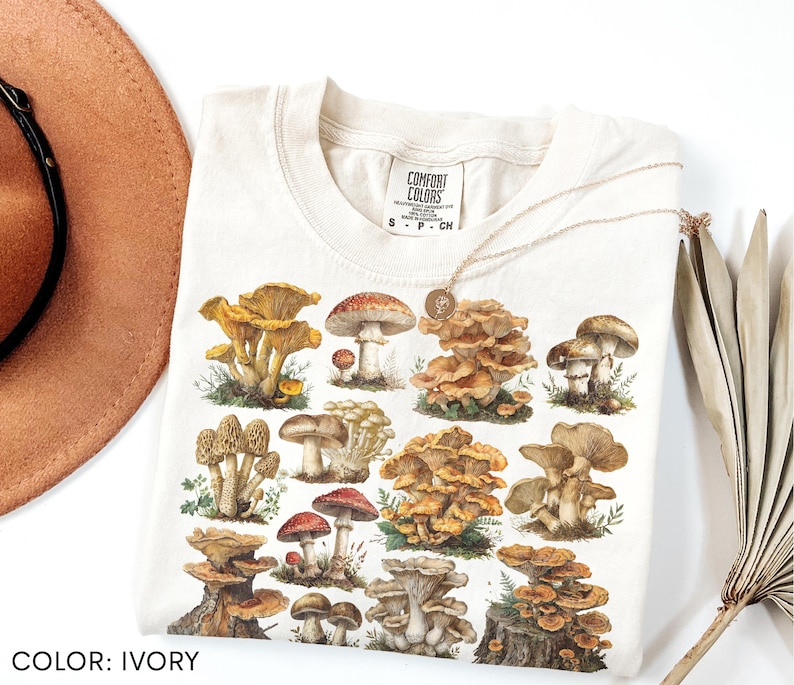 Pu&ograve; includere: T-shirt color avorio con un'illustrazione dettagliata di vari funghi nelle tonalit&agrave; del marrone, rosso e giallo. La maglietta ha un girocollo e maniche corte. Il testo "COMFORT COLORS" &egrave; stampato sulla parte superiore sinistra della maglietta.
