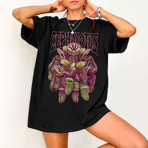 May include: A black short-sleeved t-shirt featuring a graphic print of a cluster of purple and green pitcher plants. Above the botanical illustration, the text CEPHALOTUS is printed in a stylised, dark, gothic-inspired font across the chest area.
