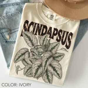 以下が含まれることがあります： アイボリー色のTシャツ。ダークな質感のフォントで「SCINDAPSUS」という文字がプリントされ、その下にハート型の葉を持つ斑入り植物のグラフィックイラストが描かれています。クルーネックで、襟の内側にComfort Colorsのラベルが付いています。