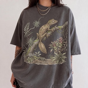 May include: A charcoal gray short-sleeve t-shirt featuring a detailed graphic print of a gecko perched on a mossy tree branch, surrounded by various tropical plants, ferns, and bromeliads in shades of green, brown, and muted earth tones.
