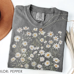 May include: A gray Comfort Colors t-shirt featuring a daisy floral design. The shirt has a crew neck and short sleeves. A silver necklace with a small pendant is draped across the shirt. The shirt is pepper colored.