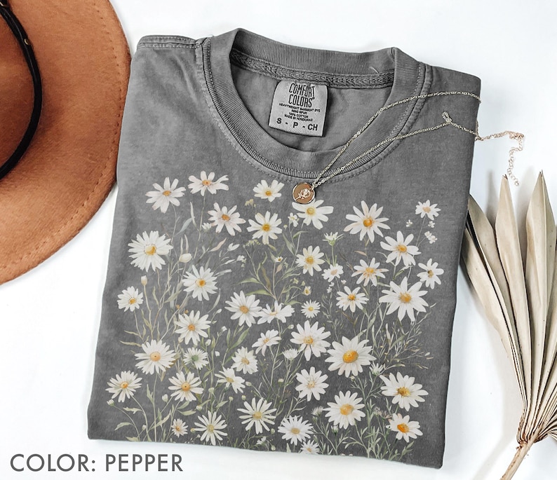 May include: A gray Comfort Colors t-shirt featuring a daisy floral design. The shirt has a crew neck and short sleeves. A silver necklace with a small pendant is draped across the shirt. The shirt is pepper colored.