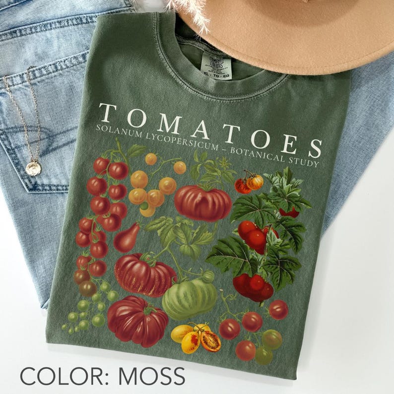 May include: Moss green t-shirt featuring a botanical illustration of various tomato varieties. The shirt displays the word "TOMATOES" in white, along with the scientific name and "BOTANICAL STUDY". The shirt is paired with blue jeans and a necklace.