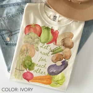 May include: Ivory t-shirt with a watercolour design of vegetables such as a tomato, pepper, and aubergine. The text "Let's root for each other" is in green script. A hat and jeans are in the background.