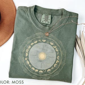 Pode incluir: Uma camiseta verde musgo com um design circular do zod&iacute;aco em dourado. A camiseta tem gola redonda e mangas curtas. Um colar de ouro com um pingente est&aacute; sobre a camiseta. O texto "COLOR: MOSS" est&aacute; na parte inferior.