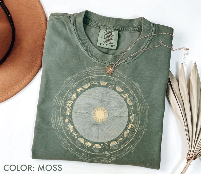 Pode incluir: Uma camiseta verde musgo com um design circular do zod&iacute;aco em dourado. A camiseta tem gola redonda e mangas curtas. Um colar de ouro com um pingente est&aacute; sobre a camiseta. O texto "COLOR: MOSS" est&aacute; na parte inferior.