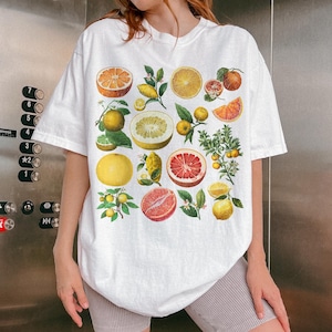 May include: A white short-sleeve t-shirt featuring a colorful graphic print of various citrus fruits, including oranges, grapefruits, lemons, and limes, displayed as whole fruits, halved sections, and small leafy branches across the front.