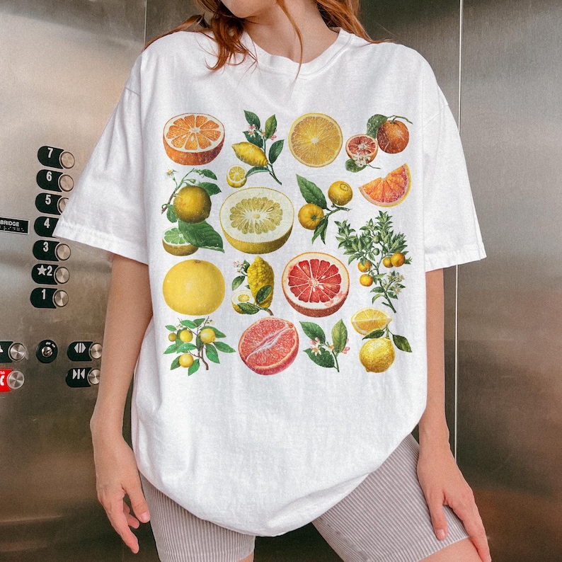 May include: A white short-sleeve t-shirt featuring a colorful graphic print of various citrus fruits, including oranges, grapefruits, lemons, and limes, displayed as whole fruits, halved sections, and small leafy branches across the front.